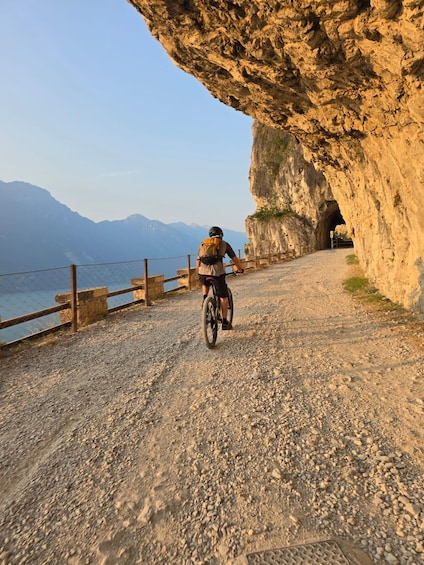Una persona montando una bicicleta de montaña sobre un camino de grava, con un acantilado rocoso a un lado y una masa de agua al fondo. 1