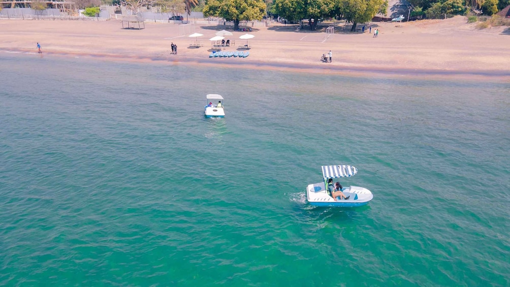 Un cuerpo de agua con dos barcos, uno con toldo a rayas y otro con toldo blanco, y una playa arenosa al fondo. 4
