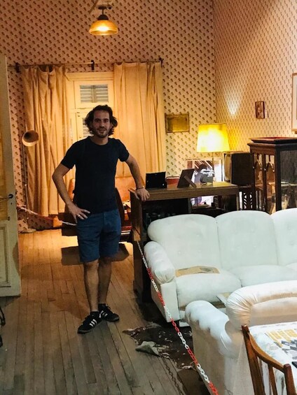Une personne se tient dans une pièce dont les murs sont recouverts d’un papier peint à motifs, avec un canapé blanc, une lampe et un ordinateur portable posé sur une table. 3
