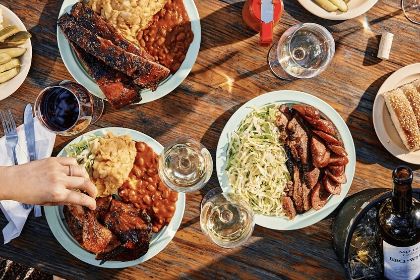 Una mesa de madera con platos de comida, incluyendo costillas, frijoles, ensalada de col y sándwiches, junto con copas de vino y una botella de cerveza. 2