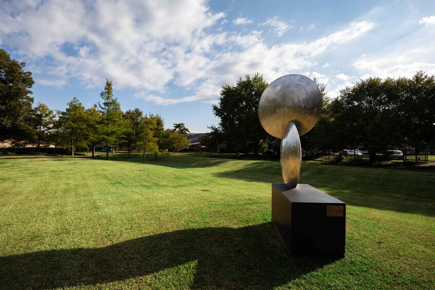 A metallic sculpture with a spherical top and a cylindrical base, set in a grassy park with trees and a clear sky. 4
