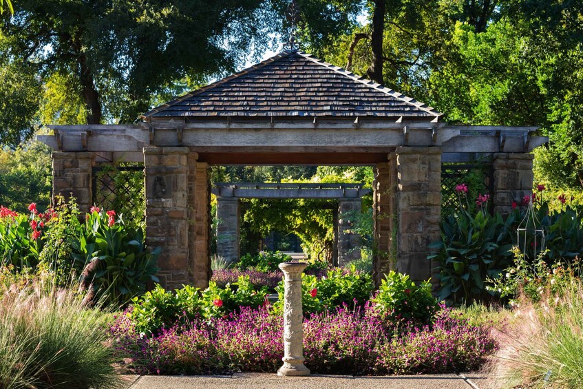A stone gazebo with a tiled roof surrounded by lush greenery and vibrant flowers. 2