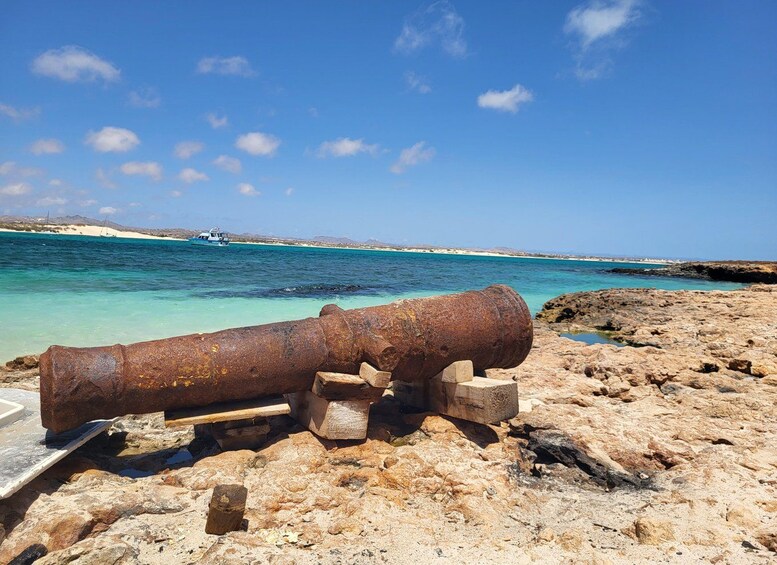 En rusten kanon på en stenet strand med klart turkis vand og en båd i det fjerne. 2