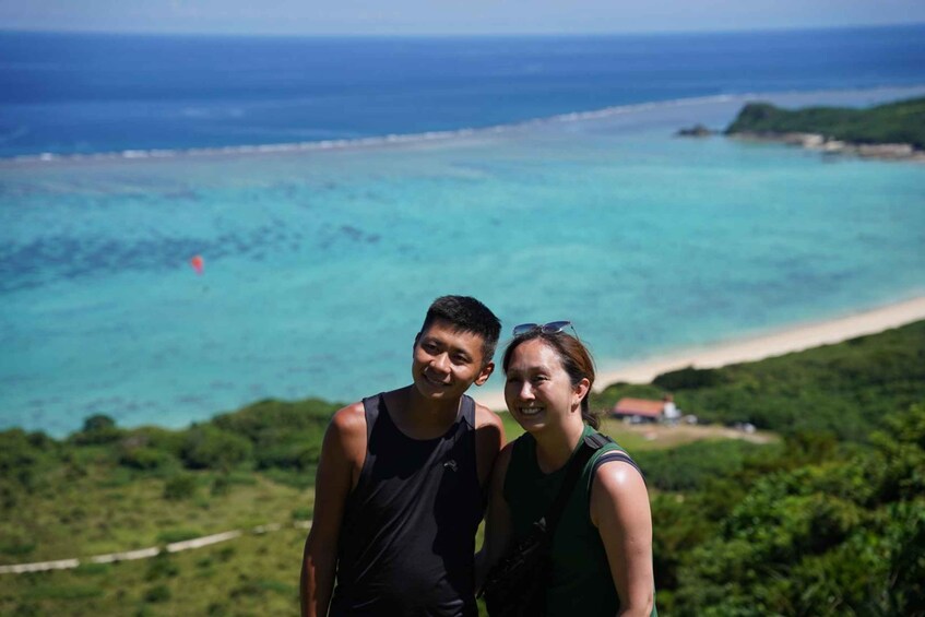 Zwei Personen stehen auf einem Hügel und blicken auf einen Strand mit klarem türkisblauem Wasser und einer entfernten Insel. 3