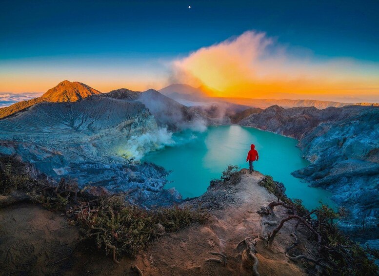 Una persona con chaqueta roja de pie sobre un afloramiento rocoso con vistas a un lago turquesa, con vapor elevándose desde el agua, rodeada de montañas escarpadas y un cielo al atardecer. 4