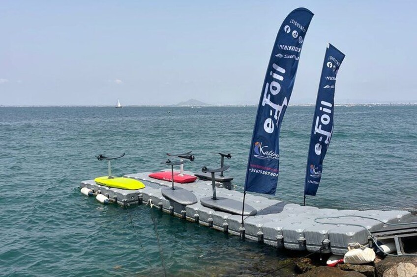A pier with kayaks and e-foil equipment, flags, and a sailboat in the background. 5