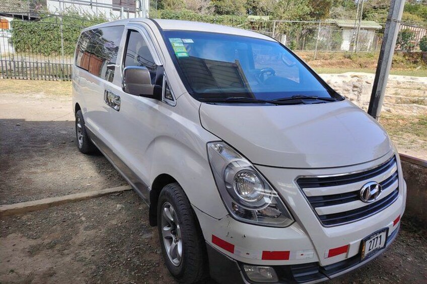 A white van parked on a gravel surface with a metal fence and greenery in the background. 4