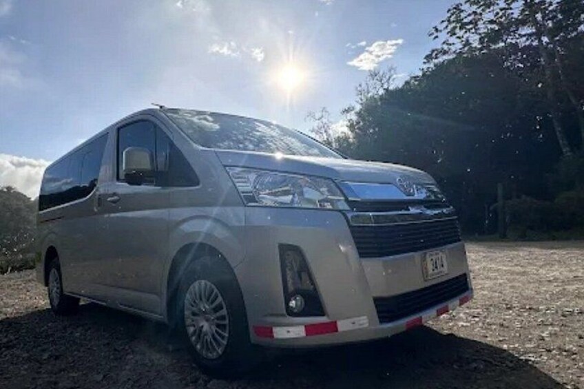 A silver minivan parked on a gravel surface with a clear sky and trees in the background. 2