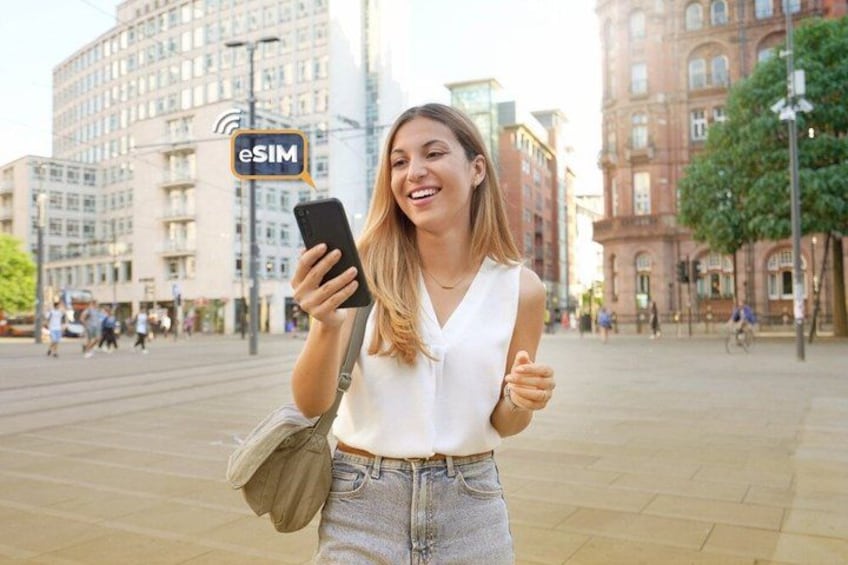 A person holding a smartphone with a visible eSIM logo, standing in an urban square with buildings in the background. 3