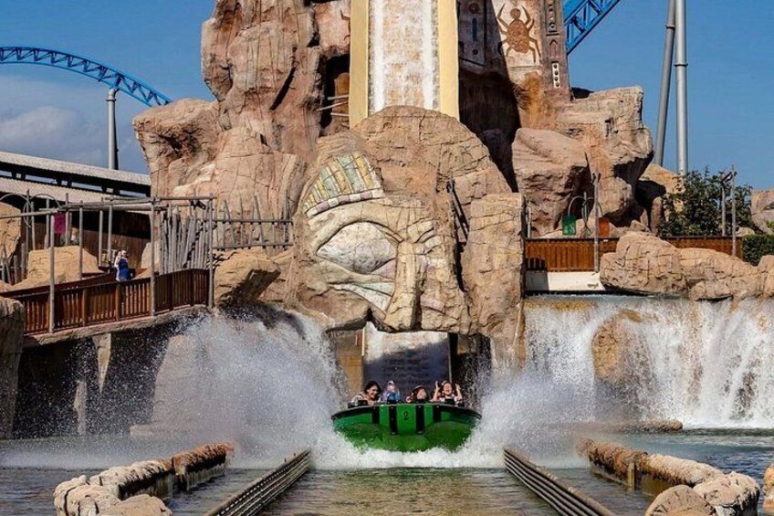 A water ride with a green boat, rocky cliffs, and a roller coaster in the background. 2