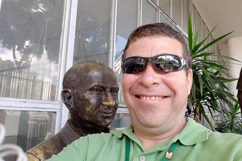 A person in a green shirt with a logo, standing in front of a window with a plant to the right. 4