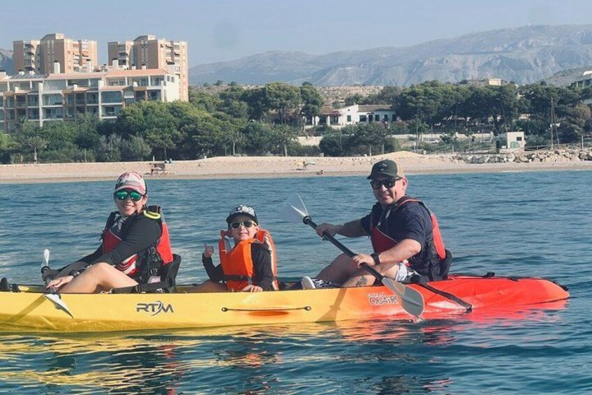 Three people kayaking on a calm body of water with a beach and buildings in the background. 2