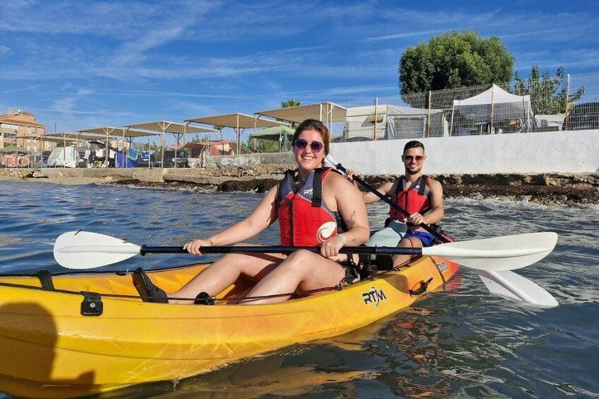 Two people kayaking on a calm body of water with a clear sky and buildings in the background. 4