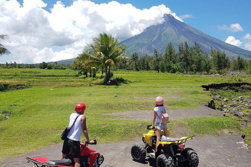 Two people on all-terrain vehicles, a volcano, and a palm tree in the background. 1