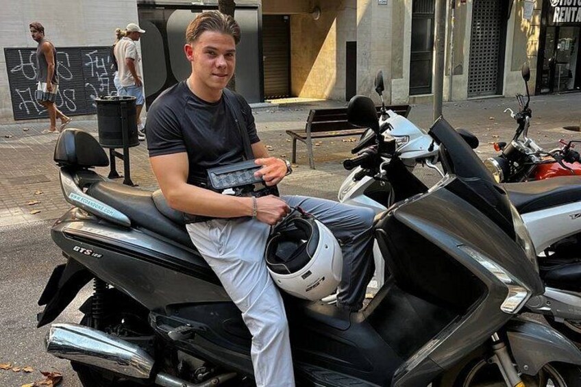 A person sitting on a black scooter with a helmet on the seat, parked on a street with buildings and a sign in the background. 1