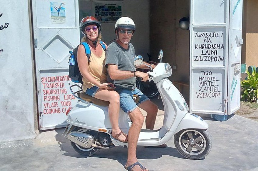 A couple on a white scooter parked in front of a building with signs for SIM Carigara Transfer, snorkeling, and village tours. 2