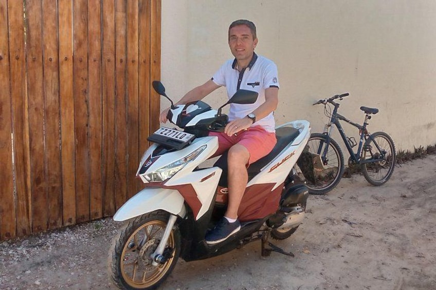 A person sitting on a scooter with a license plate, a bicycle, and a wooden fence in the background. 3