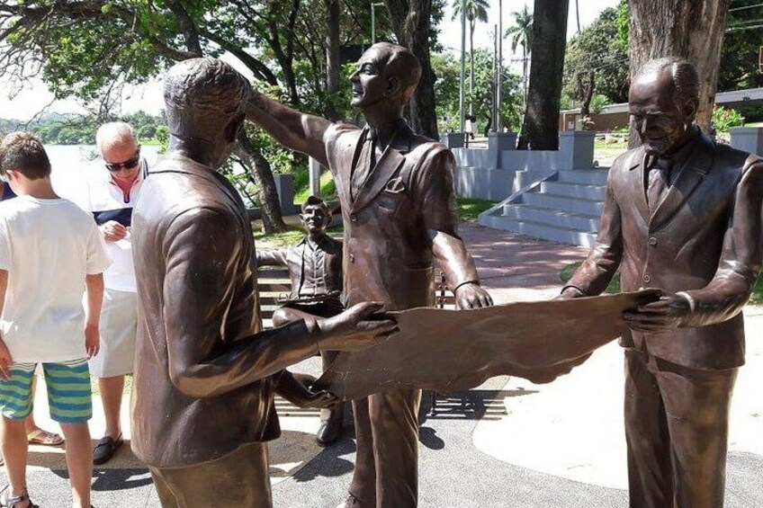A bronze statue of three figures holding a map, surrounded by trees and a paved area. 1
