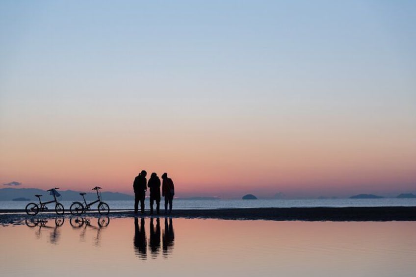 Three people standing on a beach with bicycles in the background during sunset. 1