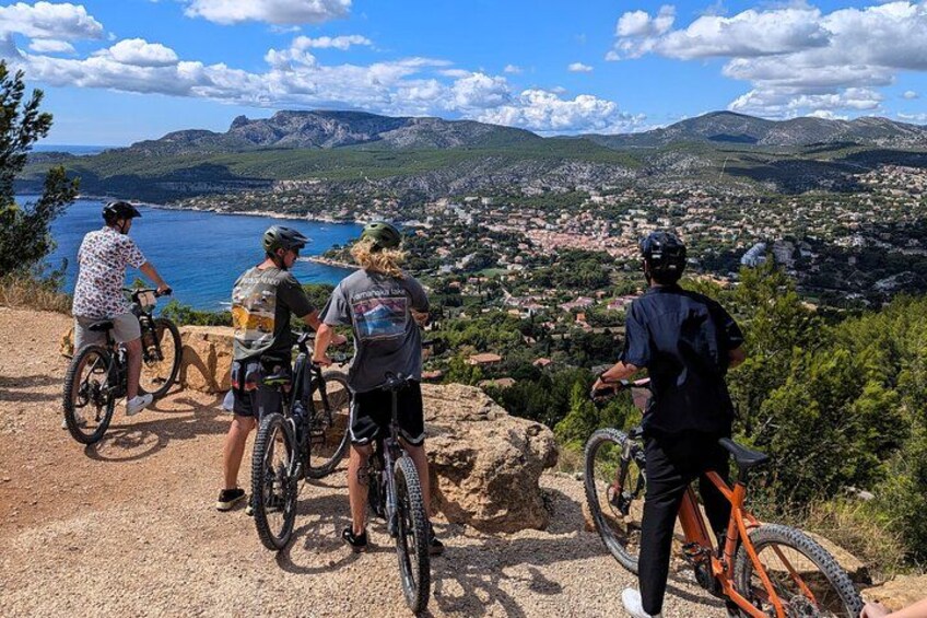 A group of mountain bikers on a dirt path overlooking a coastal town and mountains. 1