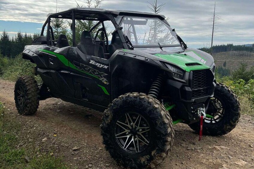 A black and green off-road vehicle parked on a dirt path with large tires, surrounded by trees and a cloudy sky. 2