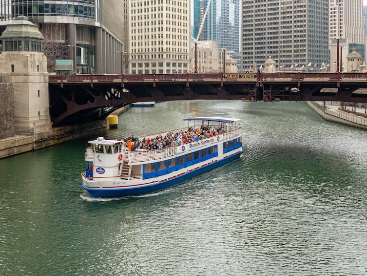 A sightseeing boat on a river with passengers, a bridge, and city buildings in the background. 1