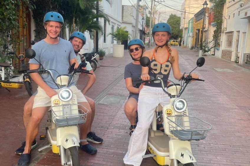 Four people on scooters, a street with buildings, and a paved road. 1