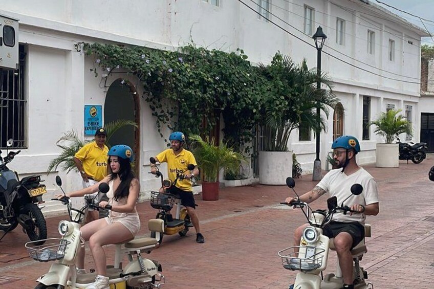 A group of people on scooters in a courtyard with white buildings and greenery. 4