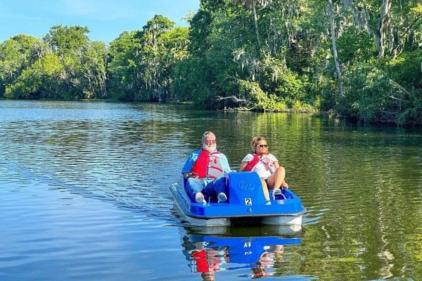 A small blue boat with two people on it, moving on calm water surrounded by dense greenery. 3