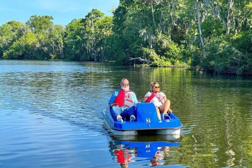 A blue boat with two people on it, moving on calm water surrounded by greenery. 3