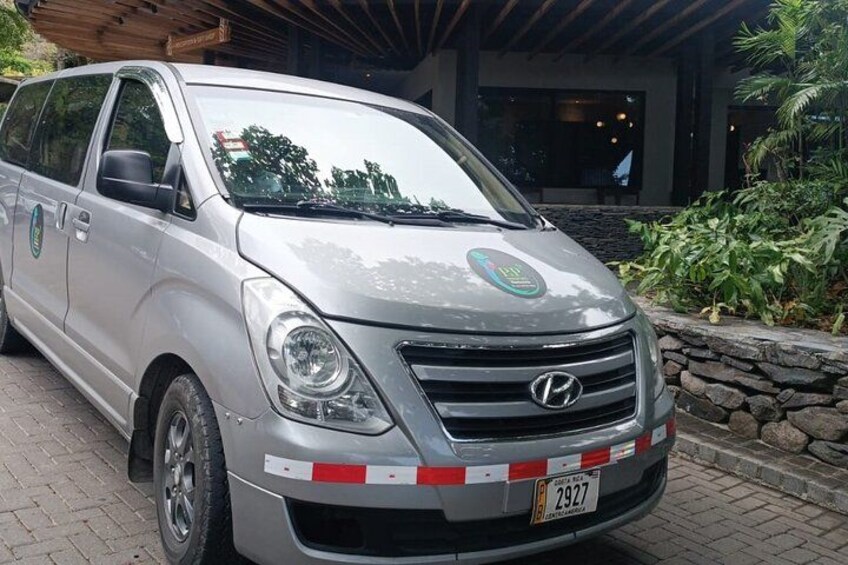 A silver van parked on a paved area with a stone wall and greenery in the background. 3