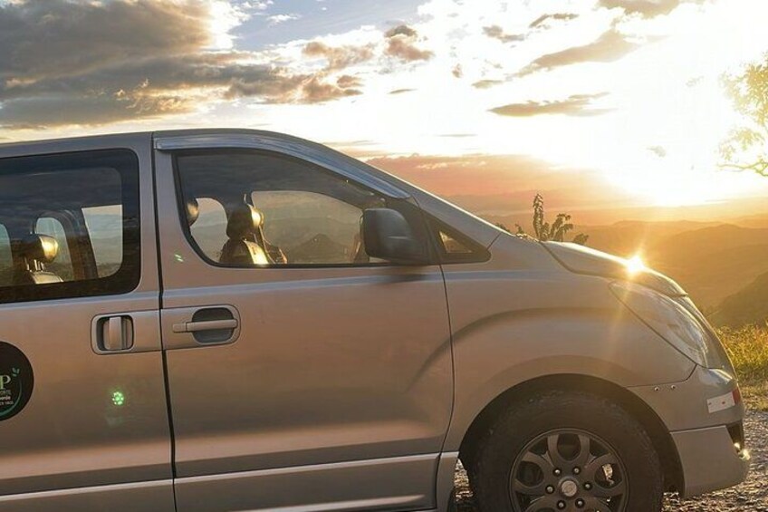 A silver car parked on a gravel surface with a sunset in the background. 1