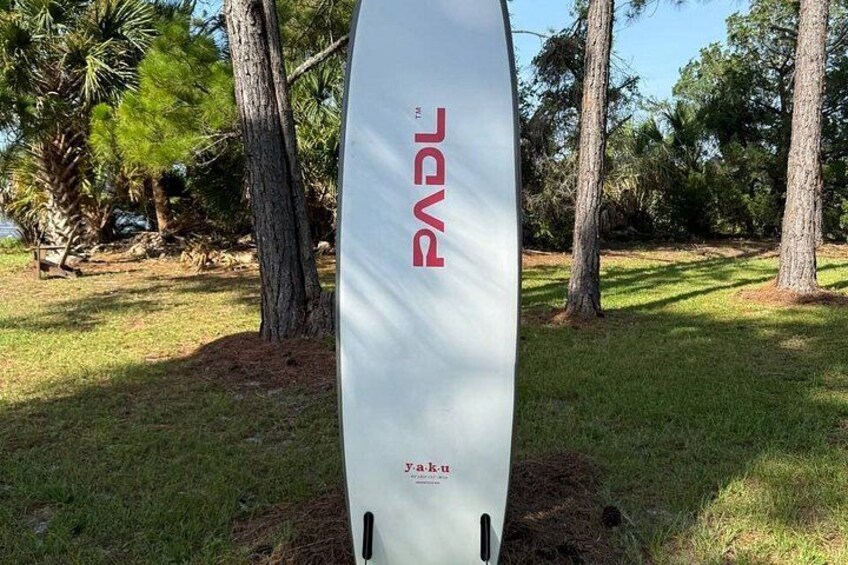 A white surfboard with the text 'PADDY' and 'Yaku' on it, standing upright in a grassy area with trees in the background. 3