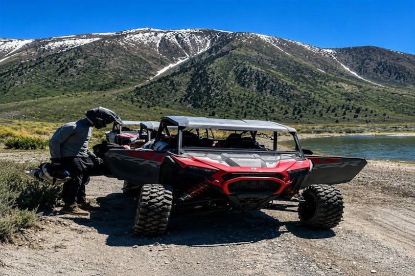 A red ATV parked on a dirt path with a person standing next to it, a lake, and mountains in the background. 2
