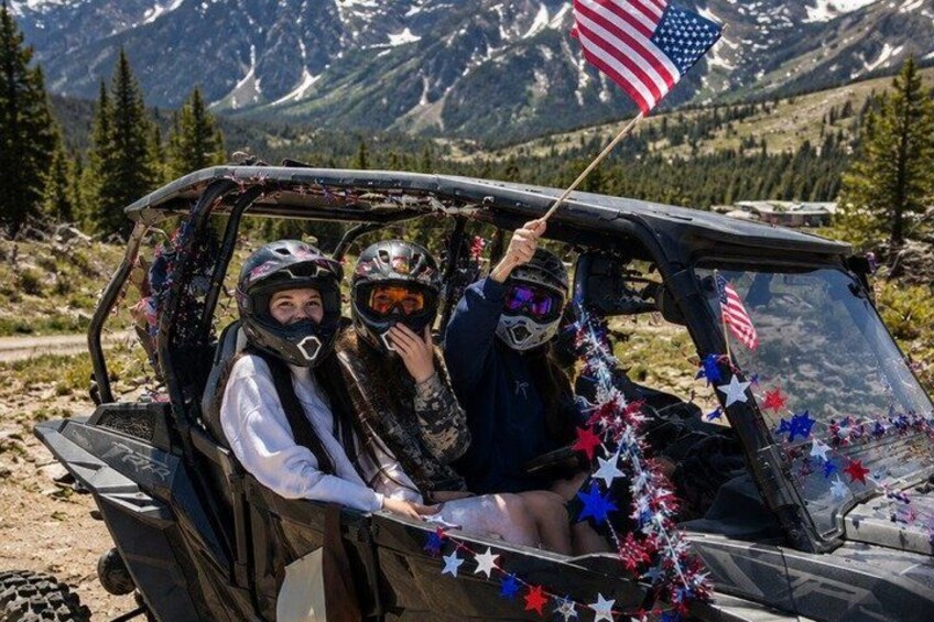 Two people in a vehicle decorated with stars and an American flag, with a mountainous landscape in the background. 5