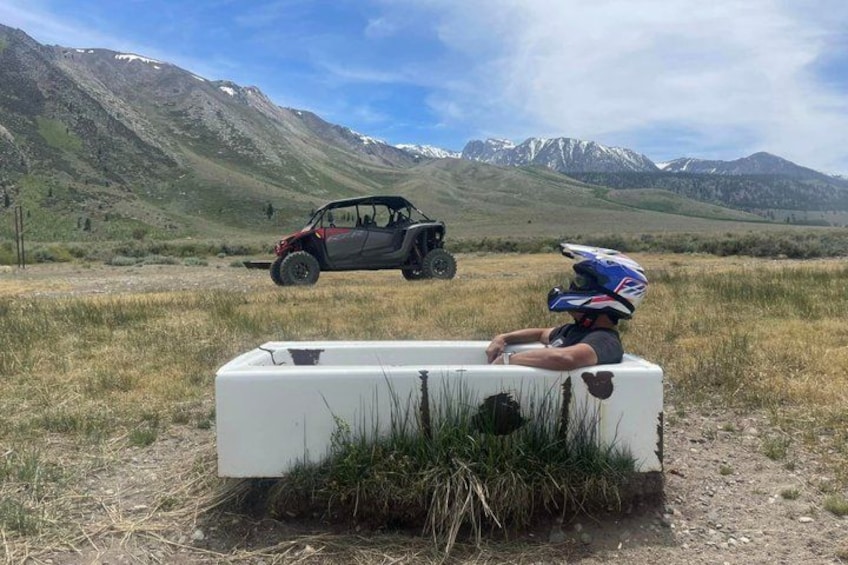 A person sitting on a white tub in a grassy field with mountains in the background and a vehicle in the distance. 1