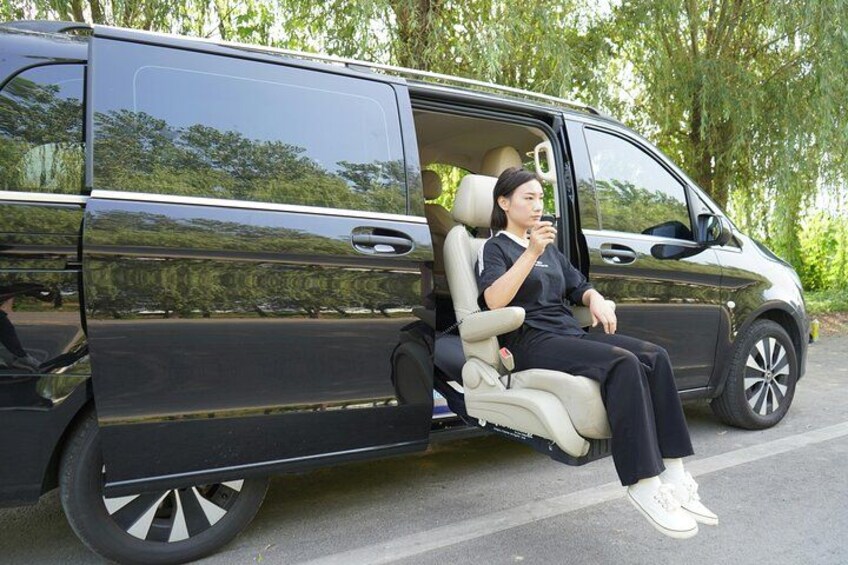 A person sitting in a black van with beige interior, parked on a road with trees in the background. 2