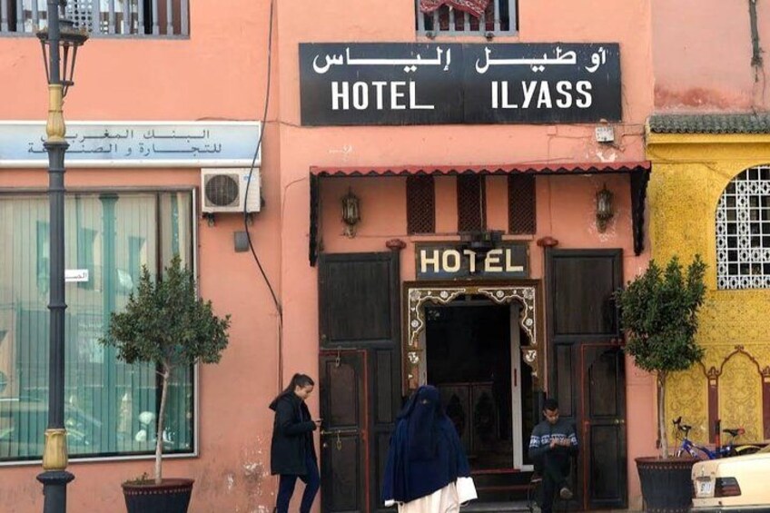 A building with a sign that reads 'HOTEL ILYASS' in Arabic and English. There are two people standing at the entrance. 5