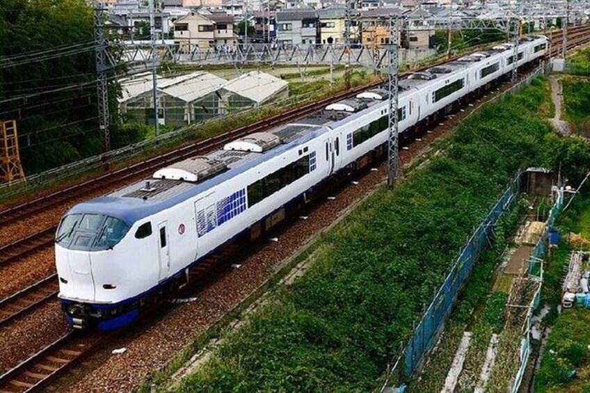 A high-speed train on tracks with overhead electrical lines, surrounded by greenery and urban buildings. 4