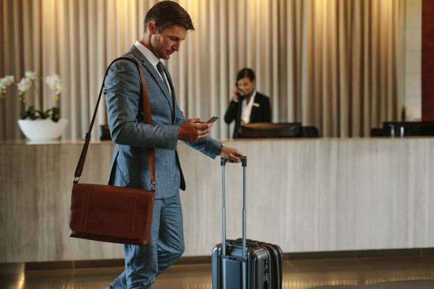 A man in a blue suit with a brown bag, using a mobile phone, standing next to a suitcase with wheels. 5