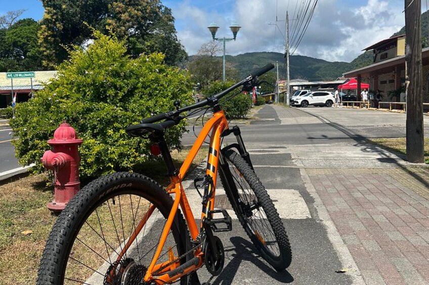 A mountain bike parked on a sidewalk next to a red fire hydrant, with a road and buildings in the background. 5