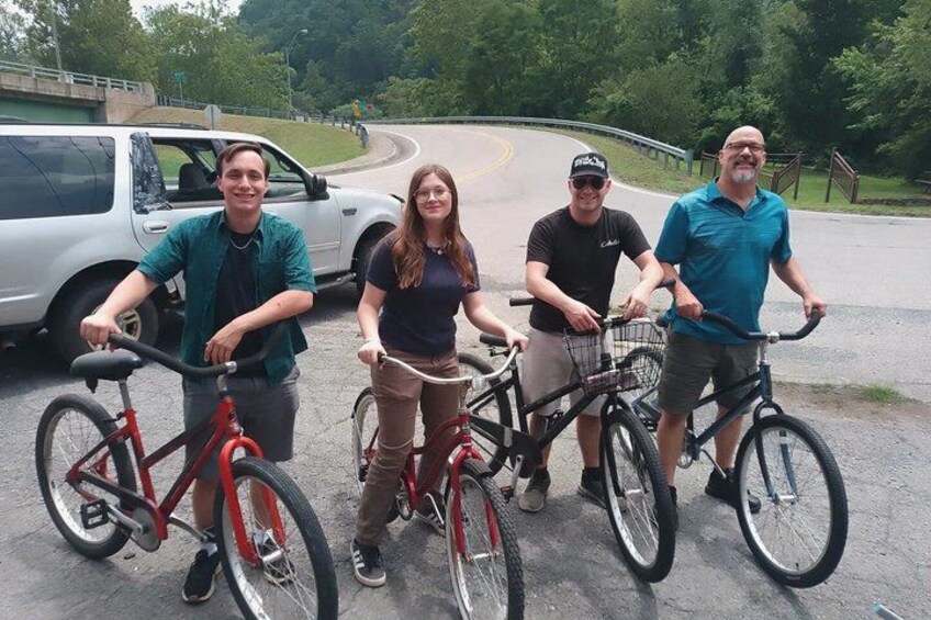 Four people standing with bicycles on a paved area, a car, and a road curving ahead. 1