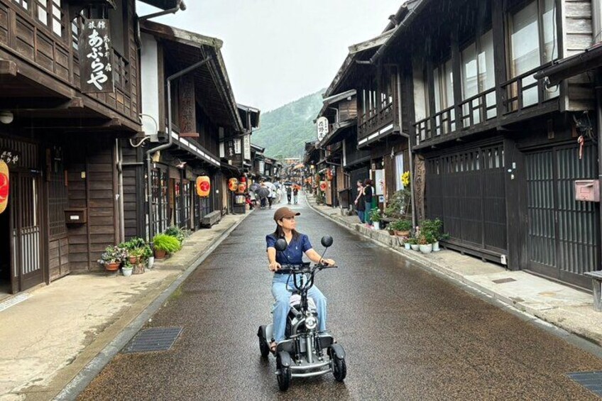 A person riding a scooter down a street lined with traditional wooden buildings. 1