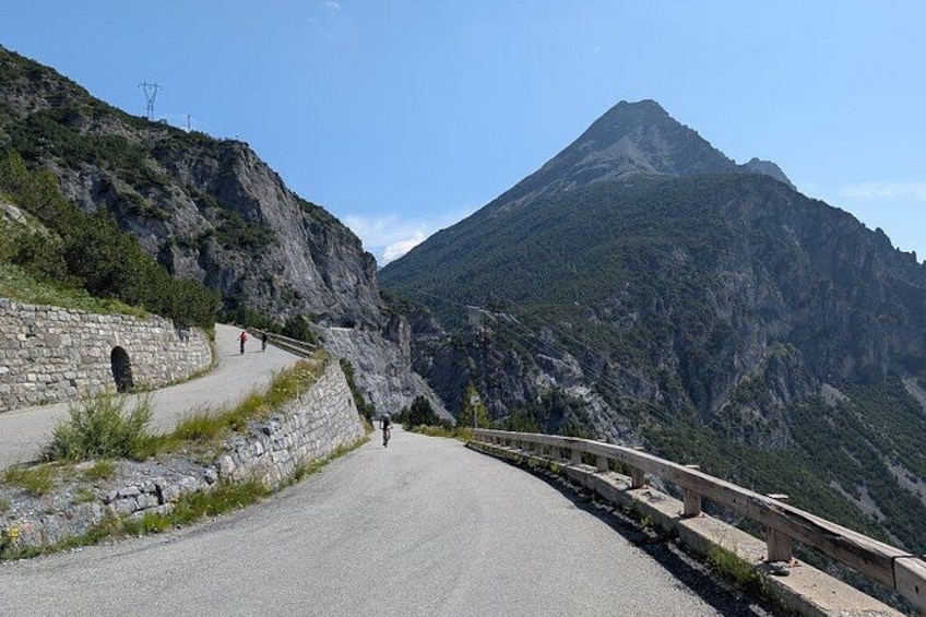 A winding mountain road with stone walls on one side and a guardrail on the other, leading towards a prominent mountain peak. 4