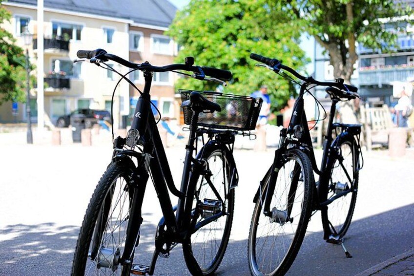 Two bicycles parked on a sidewalk with a building and trees in the background. 1