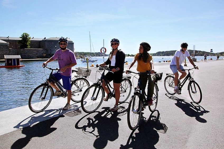 Three people on bicycles by a waterfront with a stone wall and boats in the background. 3