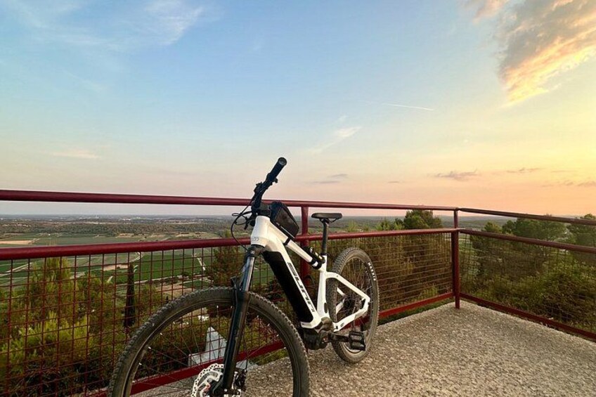 A mountain bike parked on a gravel path with a red railing, overlooking a scenic landscape with fields and a distant horizon. 2