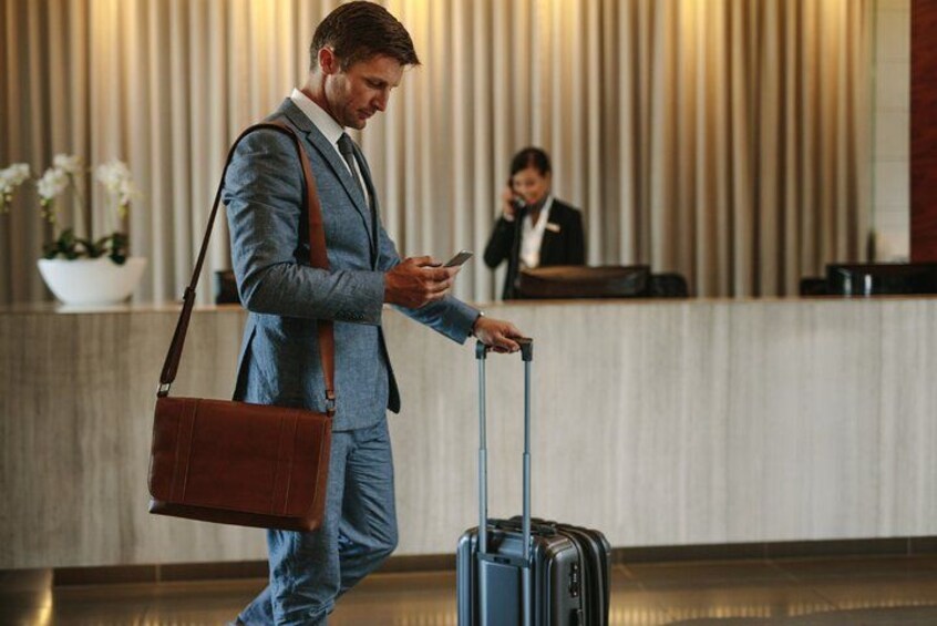 A man in a suit with a suitcase and a brown bag, standing in a lobby with a reception desk and a plant in the background. 5