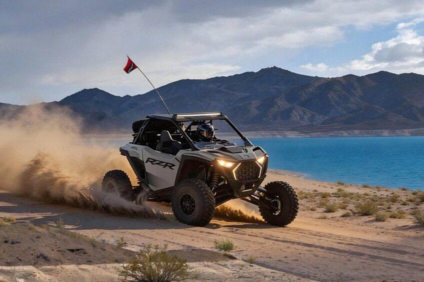 A white off-road vehicle with a red flag on the roof driving on a sandy terrain with a lake and mountains in the background. 1