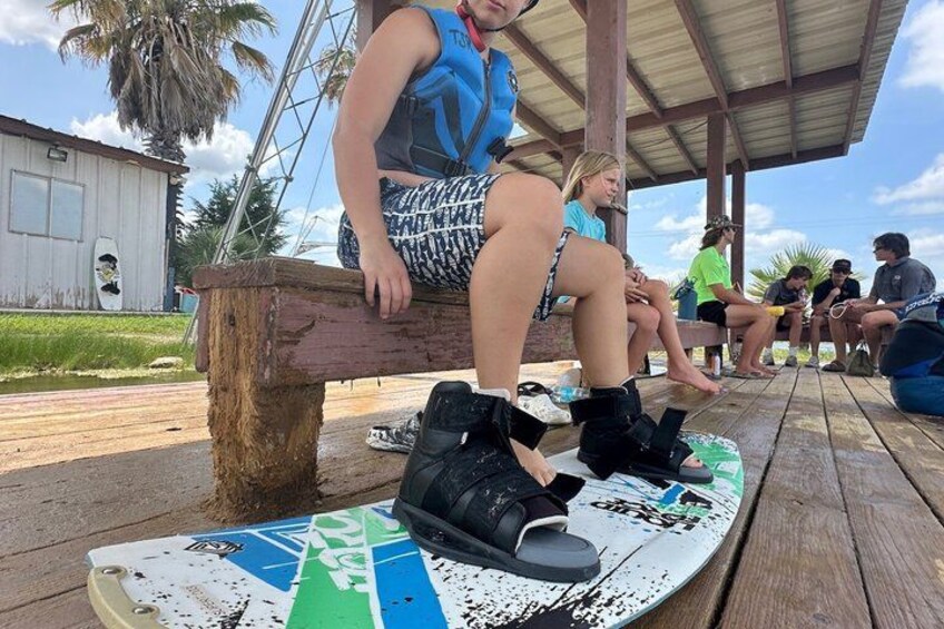 People sitting on a wooden deck, wearing water shoes, with a surfboard and a building in the background. 3